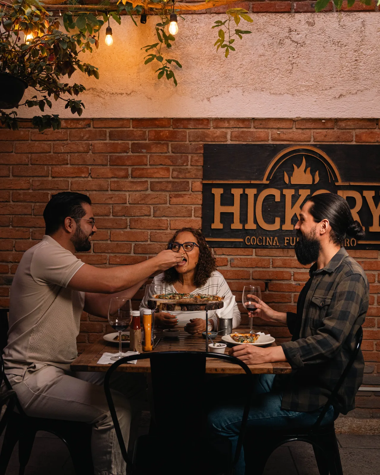 Comensales compartiendo una mesa frente al letrero de Hickory, entre ladrillo y luz cálida